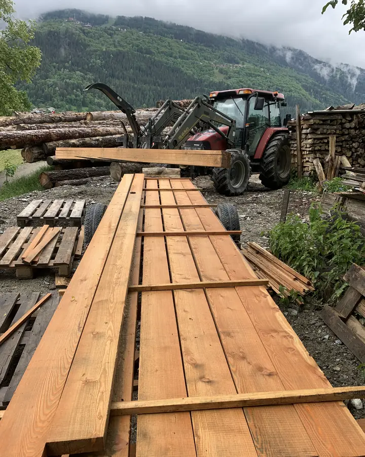 Tracteur à grappin devant des grumes, planches de mélèze fraîchement sciées au premier plan, montagne en arrière-plan